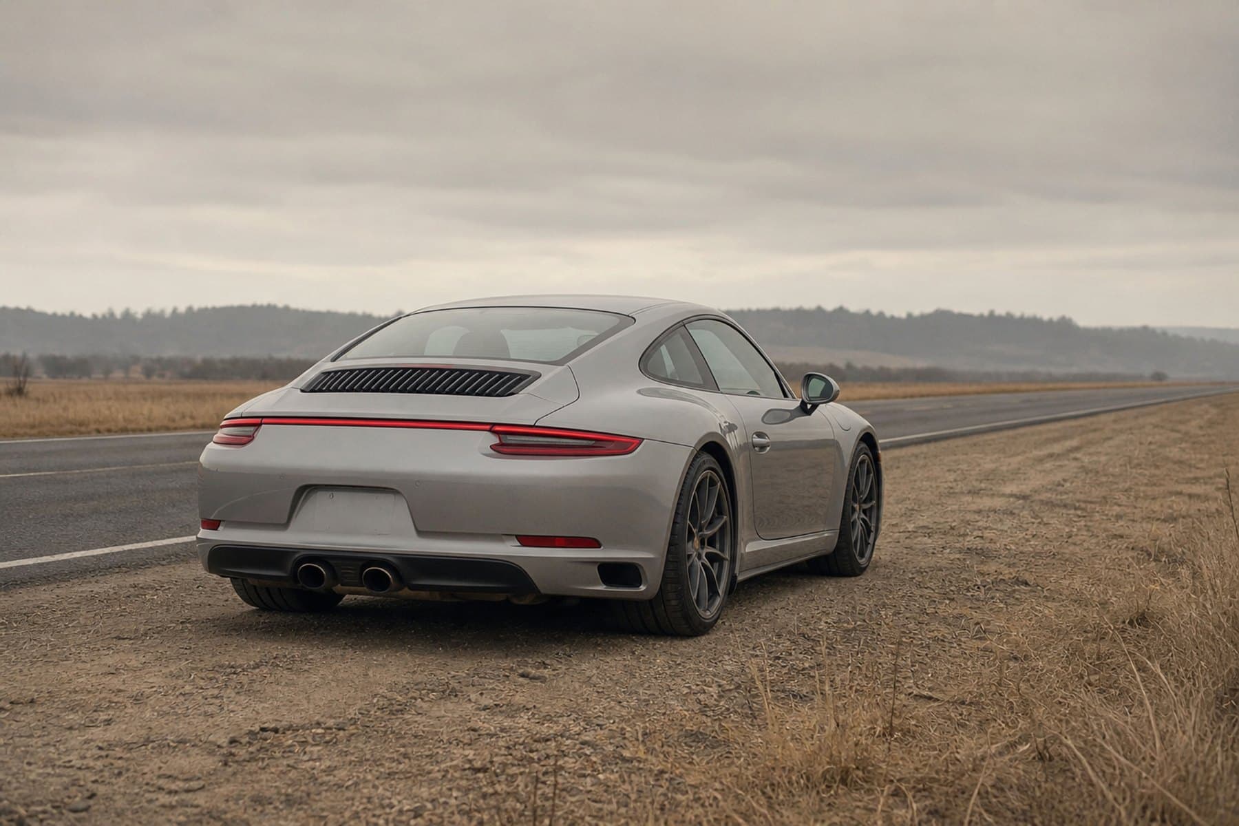 A silver Porsche 911 parked on the shoulder of an empty country road under an overcast sky
